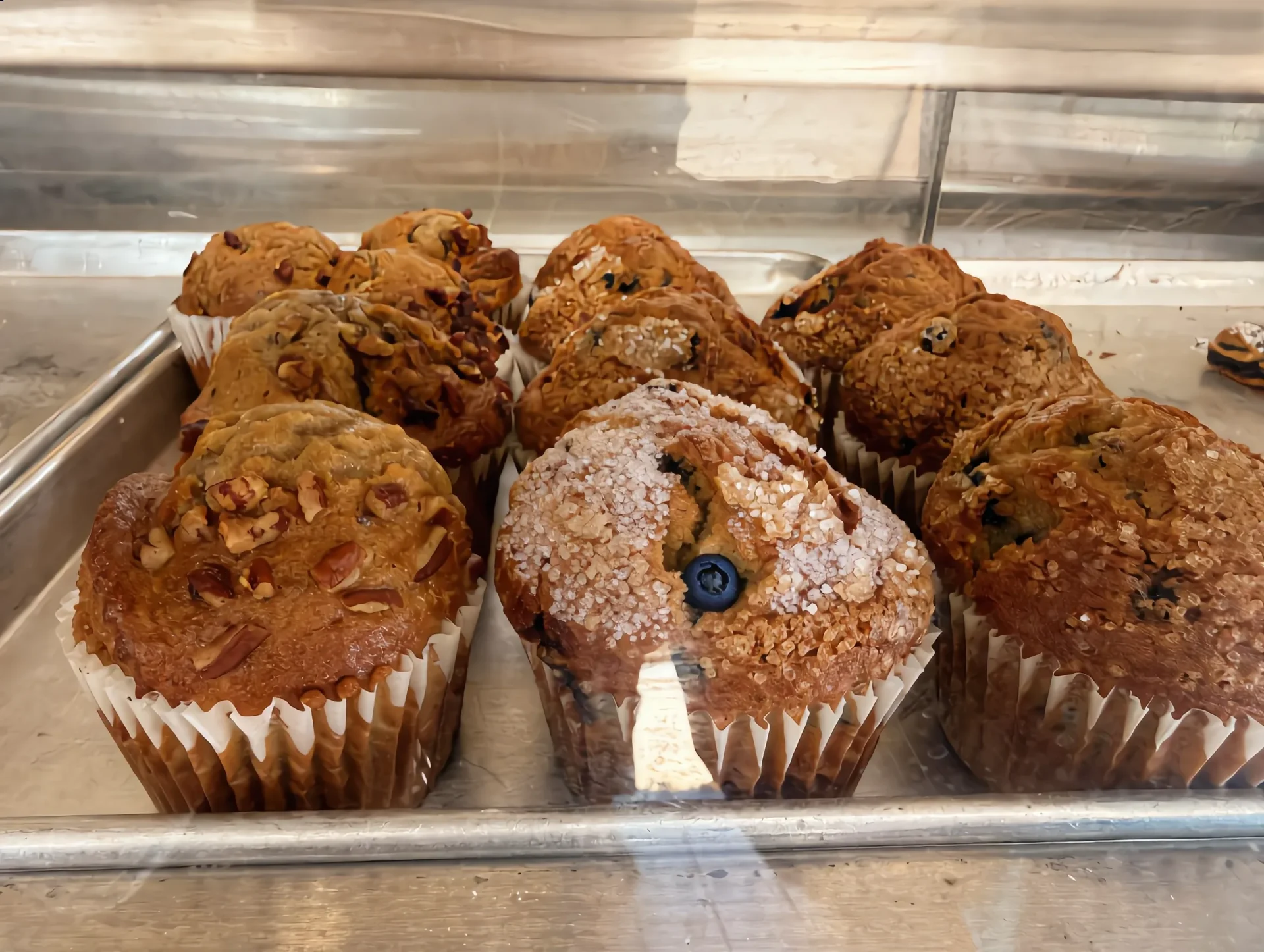 Tray of oversized bakery muffins in the display case — blueberry with sugar crust, pecan, and chocolate chip.