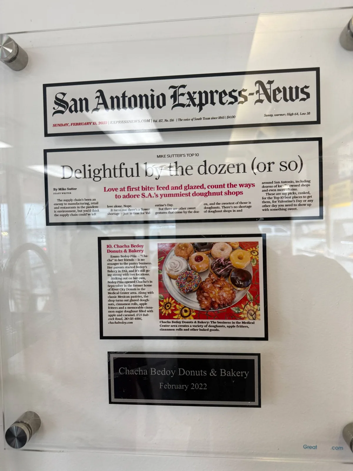 Framed San Antonio Express-News clipping from February 13, 2022 on the bakery wall — 'Delightful by the dozen (or so),' Mike Sutter's Top 10 doughnut shops in San Antonio, with ChaCha Bedoy at #10 and a colorful tray of donuts and apple fritters in the photo.